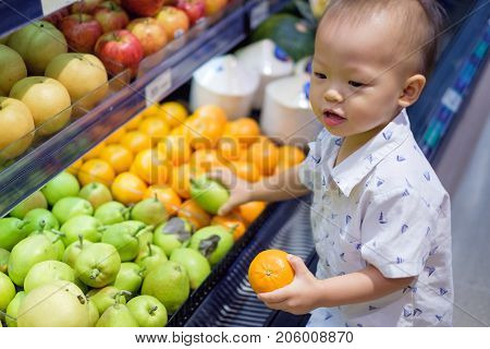 Cute little Asian 18 months / 1 year old toddler baby boy child shopping in a supermarket. Kid standing choosing and holding fruit in grocery store Kid first experience concept - Selective focus