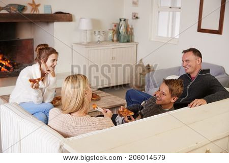 Family Sitting On Sofa In Lounge Next To Open Fire Eating Pizza