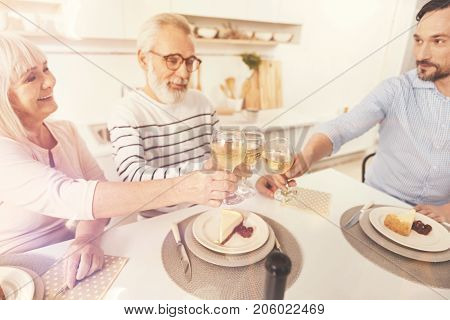 Our little tradition. Cheerful retired couple sitting at the table with their adult son while drinking wine and enjoying celebration