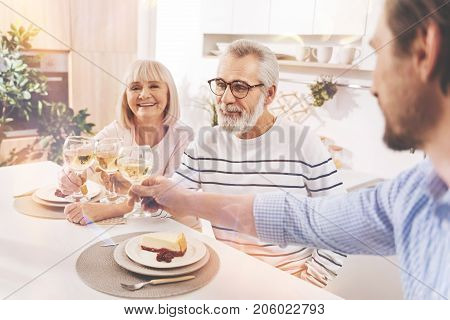 Lets make a toast. Cheerful aged couple and his son sitting in the kitchen and drinking wine while celebrating family traditions