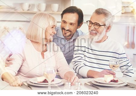 True feelings. Cheerful adult man smiling and embracing his aged parents while sitting together in the kitchen