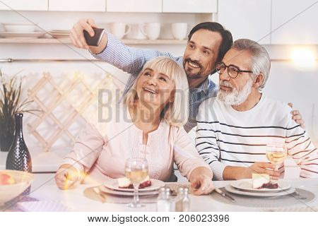 Say cheese. Overjoyed retired couple sitting in the kitchen and expressing joy while taking selfies with their adult son