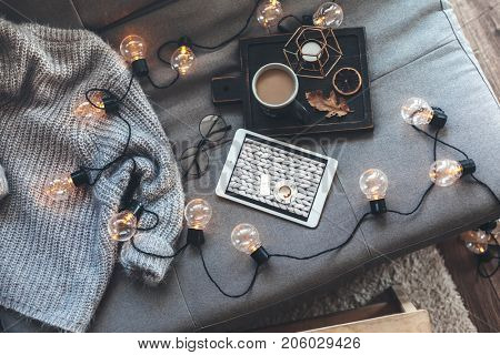 Still life details of living room. Cup of coffee on rustic wooden tray, tablet and warm woolen sweater on sofa, decorated with led lights, top view point. Autumn weekend concept. Watching movie.