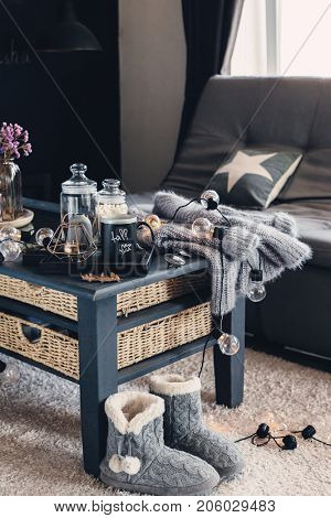 Still life details of living room interior. Cup of coffee on rustic wooden tray, candle and warm woolen sweater on table, decorated with led lights. Autumn weekend concept. Fall home decoration.