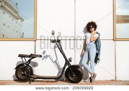 Pretty young woman in sunglasses posing while standing against a wall outdoors with a motorbike