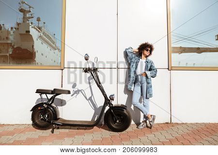 Stylish pretty woman in sunglasses posing while standing against a wall outdoors with a motorbike