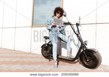 Joyful casual woman in sunglasses using tablet computer while sitting on a motorbike outdoors