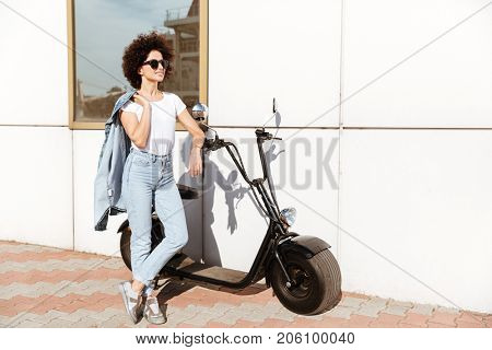 Stylish pretty woman in sunglasses posing while standing against a wall outdoors with a motorbike