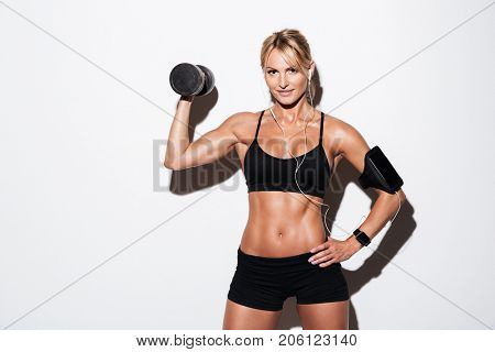 Pretty smiling sportswoman in earphones and armband standing and holding dumbbell and looking at camera isolated over white background