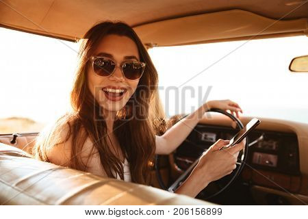 Picture from car of happy brunette woman in singlasses looking at the camera while holding smartphone in hand