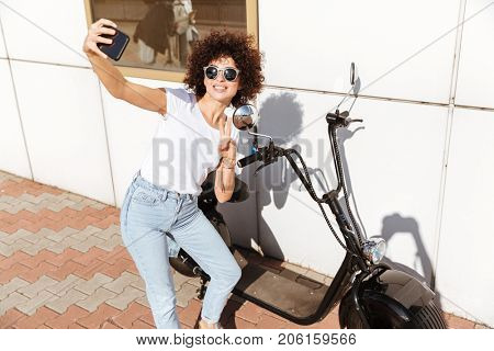 Cheerful attractive girl in sunglasses taking a selfie and showing victory gesture while leaning on a motorbike outdoors