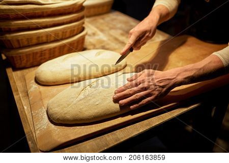 food cooking, baking and people concept - baker hands making bread and cutting dough with knife at bakery