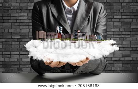 Cropped image of businessman in suit presenting in his hands modern city block with gray brick wall on background. 3D rendering.