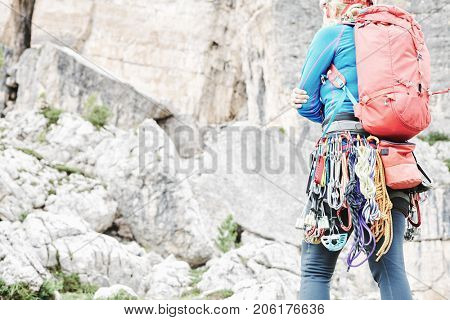 Female mountaineer with trad climbing rack including backpack, helmet, chalk bag, harness with spring-loaded cams, nuts, quickdraws, slings and carabiners preparing for ascent in summer Dolomite Alps