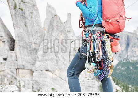 Close-up of mountaineer with trad climbing rack including backpack, chalk bag, harness with spring-loaded cams, nuts, quickdraws, slings and carabiners preparing for ascent in summer mountains