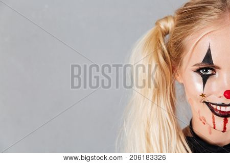 Half face portrait of a smiling blond woman in halloween clown make-up and blood streaks on her face looking at camera isolated over gray background