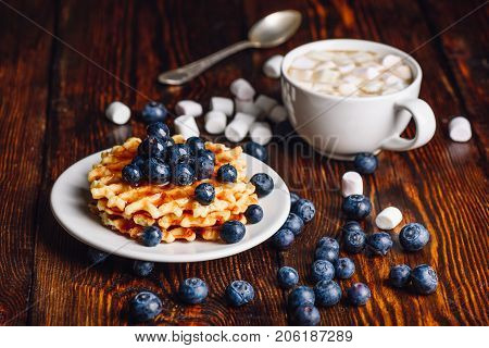 Belgian Waffles on Plate with Fresh Blueberry and Cup of Hot Chocolate with Marshmallow. Spoon and Some Berries and Marshmallows Scattered on Wooden Background.