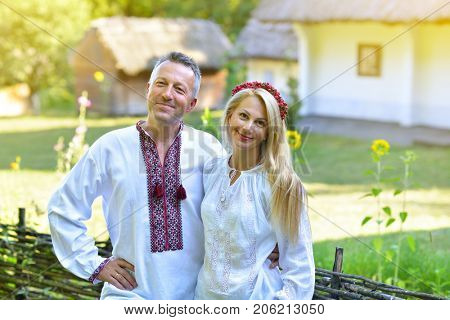 Ukrainian couple in traditional clothing vyshyvanka with embroidery over landscape of tipical ucrainian village with old houses, summer outdoor. Ukrainian people posing near the family house
