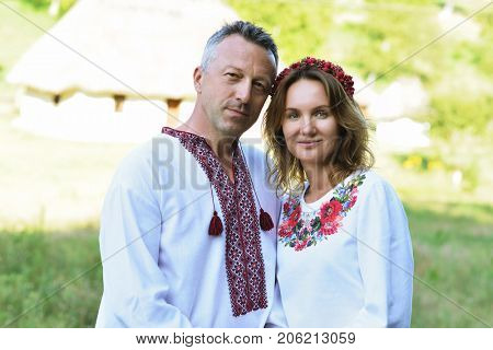 Ukrainian couple in traditional clothing vyshyvanka over landscape of tipical ucrainian village, summer outdoor. Ukrainian people posing near the house