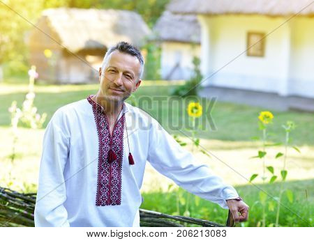 Ukrainian man in traditional clothing vyshyvanka with embroidery over landscape of tipical ukrainian village with old houses, summer outdoor.