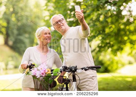 active old age, technology and lifestyle concept - happy senior couple walking with fixie bicycles taking selfie by smartphone at summer park