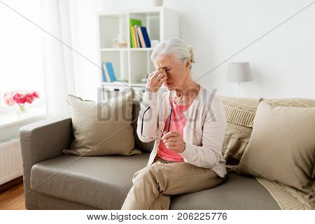old age, health problem, vision and people concept - senior woman with glasses sitting on sofa and having headache at home