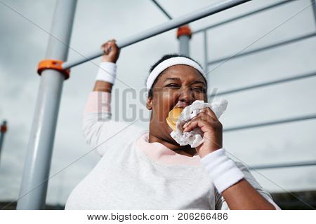 Very hungry young overweight woman eating sandwich while holding by bar of outdoor sport facilities during physical exercise