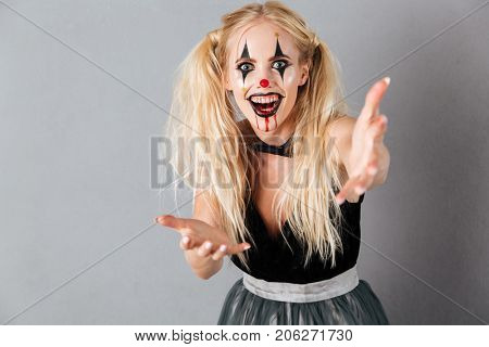 Happy blonde woman in halloween make up extends her hands and looking at the camera with open mouth over gray background