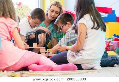 Cute pre-school boy cooperating with his colleagues at the construction of a structure made of wooden toy blocks, under the guidance of a young kindergarten teacher