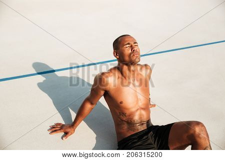 Close up portrait of a half naked muscular afro american sportsman resting while sitting on a ground at the track field