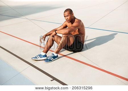 Half naked young african sportsman resting while sitting on a ground at the track field with water bottle