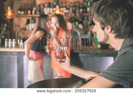 Handsome drunk man sitting at bar, drinking cocktail and looking at two attractive girls. He wants to get to know them, but does not know how. Nightclub. Date