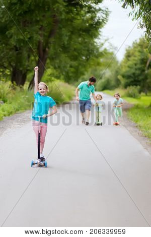 Father teaching his cute little daughters to ride kick scooter at the park on a sunny summer day.