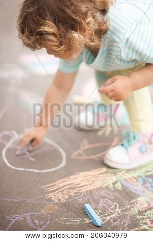 Sunny and warm summer days and happy chilhood. Cute little girl drawing with chalk crayons at the asphalt.