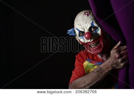 closeup of a scary evil clown peering out from a purple stage curtain, with a blank black space on the left
