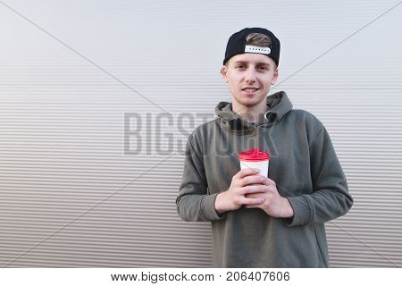 Portrait of a stylish young student in a cap on a light background with a cup of hot drink in her hands.