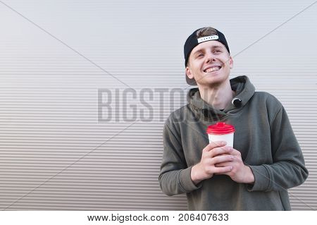 Portrait of a positive smiling student in a cap on a light background with a cup of hot drink in his hands.