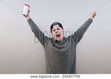 Happy young man with coffee in the hands of happiness lifted his hands upward against the background of a light wall