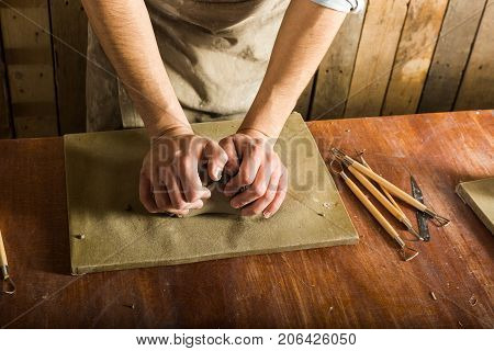 potter, workshop, ceramics art concept - closeup on male hands kneading the fireclay, a ceramist with raw material on wooden table with sculpting tool set, top view