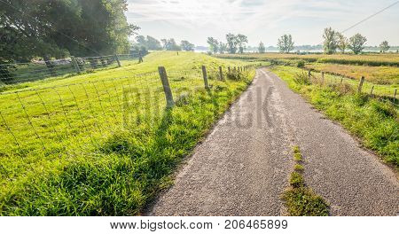 Backlit image of a narrow country road between fences. It is early in the morning of a sunny day in the beginning of the fall season. In the background is a wide Dutch river.