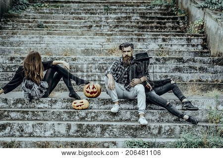 Halloween bearded man sitting with girls. Halloween jack o lantern symbols. Holiday celebration concept. People or friends on stony stairs. Women with pumpkin heads.
