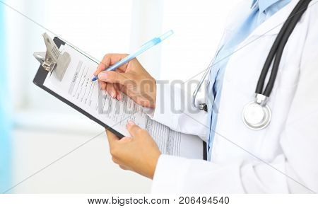 Close-up of a female doctor filling  up medical form at clipboard while standing straight in hospital.