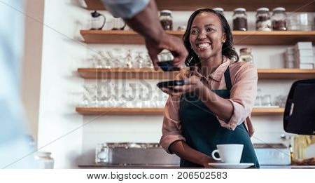 Smiling barista using nfs technology to help a customer pay for a purchase with their bank card in a cafe
