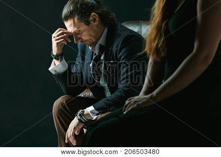 The actor or mature man wearing suit in a classical retro fashion style. The stylish man sitting on a leather couch in a studio. The woman standing near