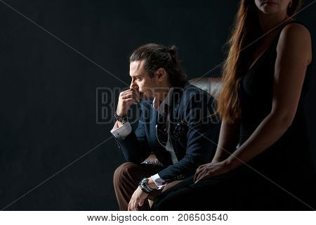 The actor or mature man wearing suit in a classical retro fashion style. The stylish man sitting on a leather couch in a studio. The woman standing near
