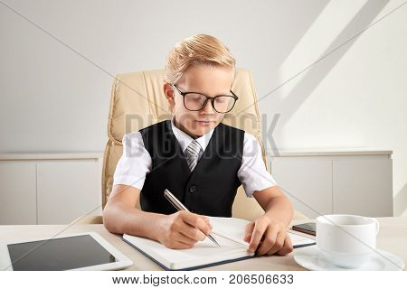 Serious little business executive in glasses writing ideas into textbok