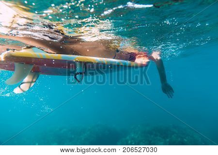 Young girl in bikini - surfer with red surf board in underwater. Ripples at water surface with sky color.