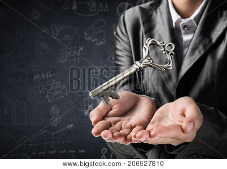 Cropped image of businessman in suit keeping big key in hands with business sketches on dark wall on background.