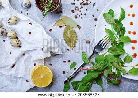 A white plate with drops of sauce and fresh green leaves, bay leaves, a half of juicy yellow lemon, little speckled quail eggs, a jar with different seasonings on a light gray background, top view.