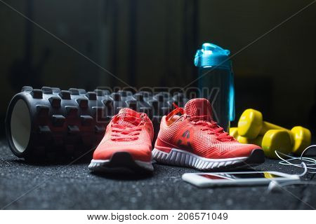A blue bottle for water, yellow dumbbells, a white mobile phone with headphones and pink vivid sheakers for sport routine on a dark blurred background.